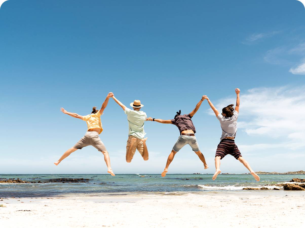 Foto tirada com a câmera Ultra Wide de quatro homens de mãos dadas pulando em uma praia durante o dia.  Uma visão mais ampla da praia abaixo e do céu é visível.